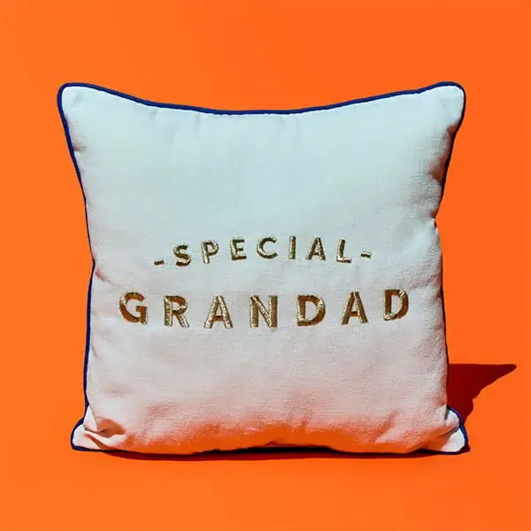 A white cushion with gold embroidered text reading Special Grandad, displayed against a bright orange background.